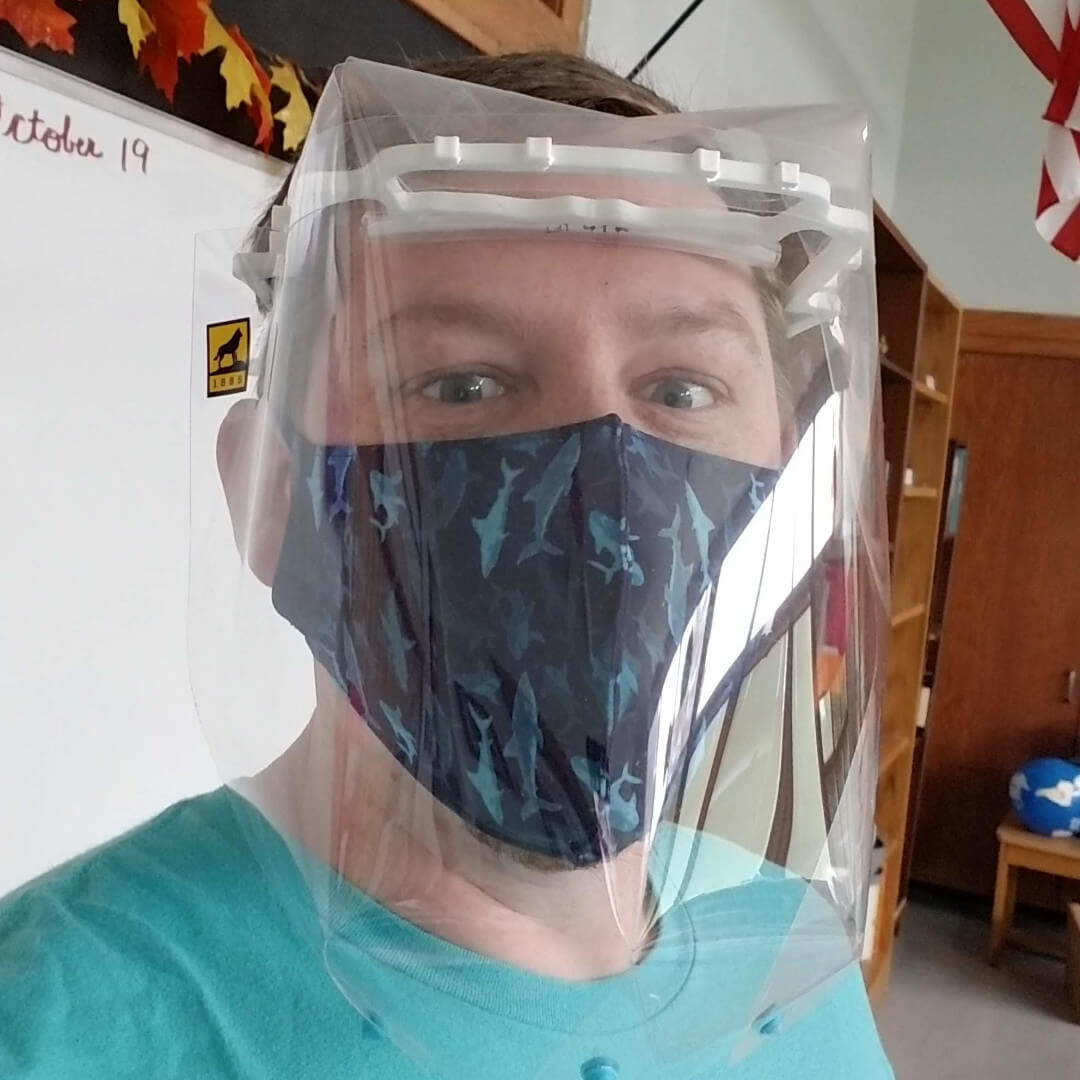 Man with face shield and mask in front of whiteboard.